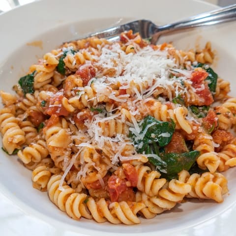 A close-up of Tomato Spinach One-Pot Rotini garnished with grated Parmesan, served alongside a slice of warm crusty bread for dipping.  