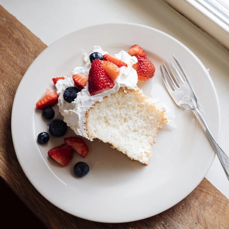 A slice of angel food cake on a white plate, topped with fresh strawberries and a generous dollop of whipped cream.